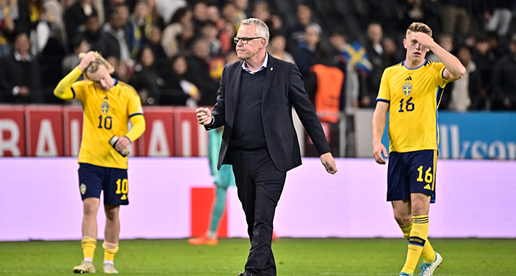 Man i kostym mellan två fotbollsspelare på en fotbollsplan.