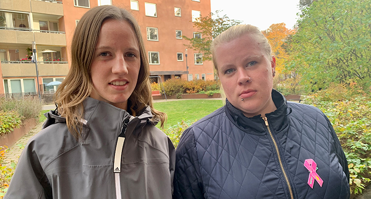 De står bredvid varandra. De tittar in i kameran. Bakom syns hus och träd.