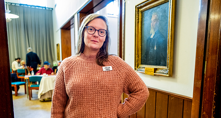 Hon står framför en tavla med armen i sidan. Bakom syns bord där människor står och sitter