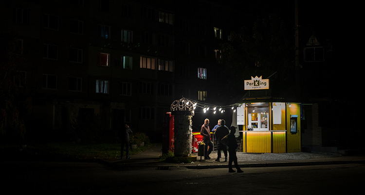 Ett litet hus är upplyst av några lampor, men det är svart runtomkring. I ett hyreshus bakom är det mörkt med bara några få svaga lampor i fönster.