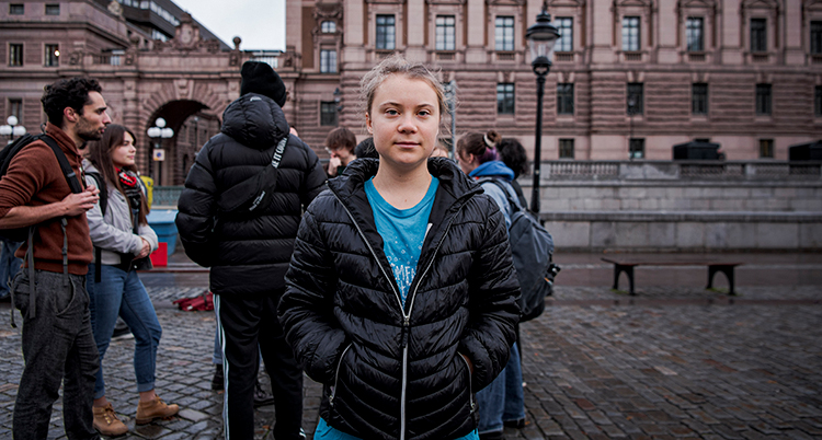 Hon står framför riksdagshuset. Hon har jacka och håller händerna i fickorna. Hon tottar rakt in i kameran. Bakom syns andra människor.