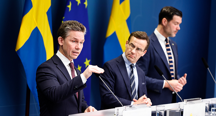 Tre män står bredvid varandra i talarstolar. De har kostymer. Bakom syns svenska flaggor.