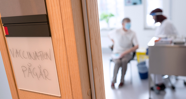 En person får en spruta i ett rum, bilden på personerna är suddig. I förgrunden syns en dörrkarm där det står en skylt: vaccination pågår.