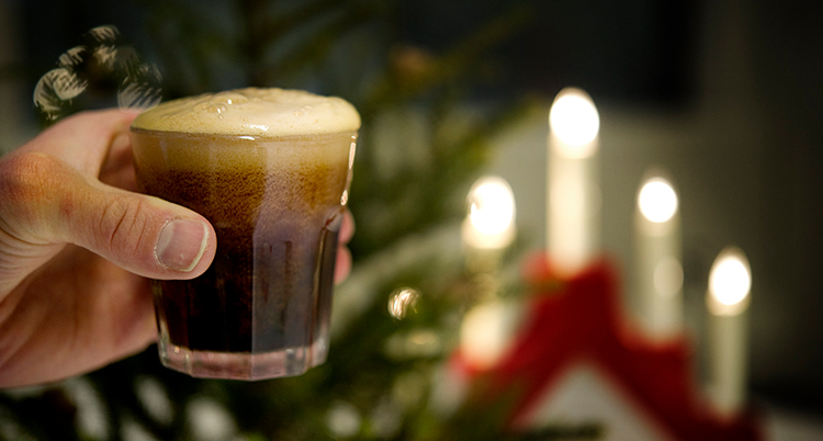 En hand håller i ett glas med brun bubblande dryck. Bakom syns en elljusstake