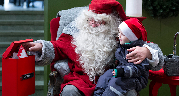 En tomte med stort vitt skägg, röd luva och röda kläder har ett barn i knät. Tomten sitte ri en fåtölj och sträcker ut en hand och öppnar en röd brevlåda med post i.
