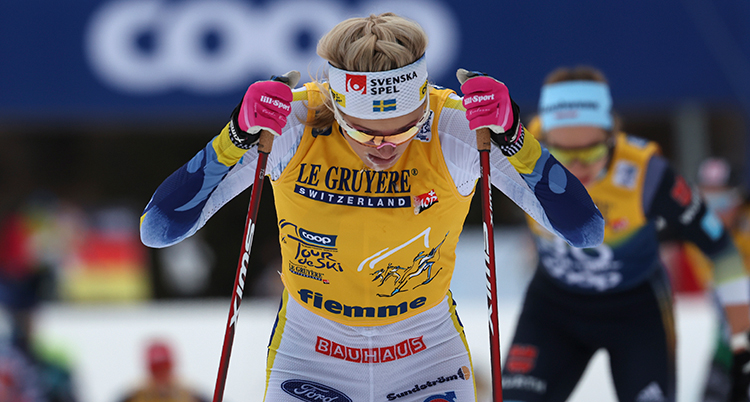 Hon stakar med stavarna och tittar ner. Hon har vit skiddräkt och gul nummerlapp.