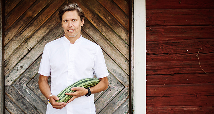 Han står utanför ett hus och tittar in i kameran. Han håller i en stor zucchini.