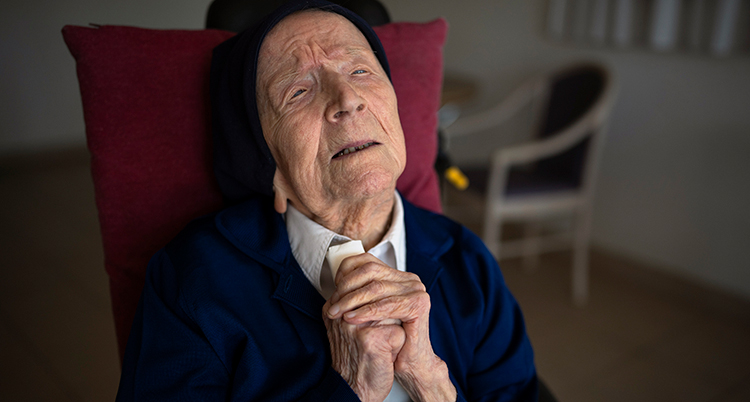 Hon lutar huvudet bakåt och blundar. Hon håller händerna knäppta framför sig. Hon sitter i en röd fåtölj i ett rum.