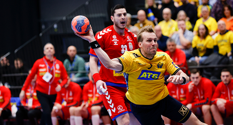 Han har blågula sportkläder. Han är på väg att kasta iväg bollen. Bakom syns rödklädda spelare.