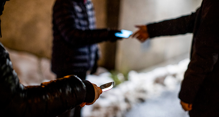 En hand som håller i en kniv syns längst fram i bilden. Bakom syns två personer med jackor. Den ena personen sträcker en mobiltelefon till den andra.