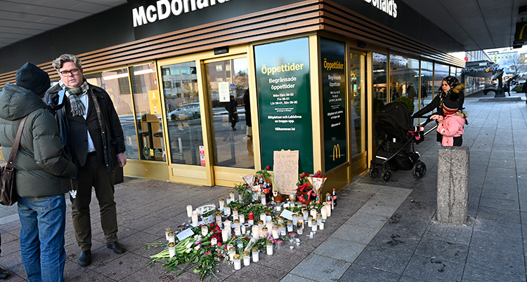 Han står till vänster i bild och blir intervjuad av en person i jacka. Han står utomhus, framför en restaurang. På marken finns blommor och ljus. En kvinna med barnvagn går in i restaurangen till höger i bild.