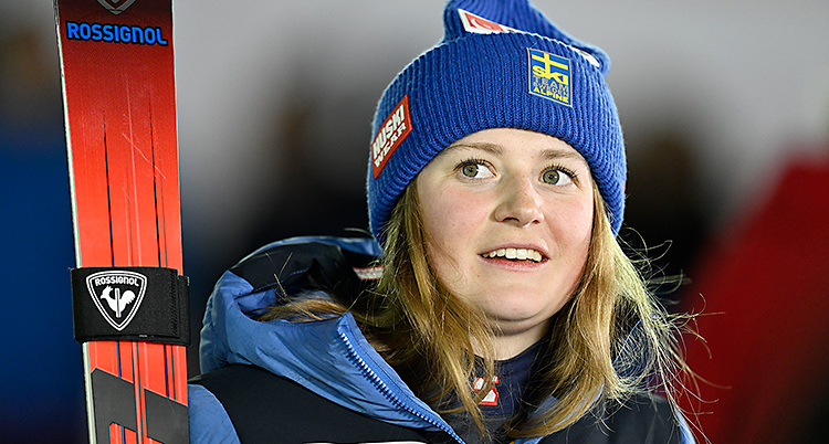 Hon står och håller i sina skidor. Hon har en blå mössa på huvudet.
