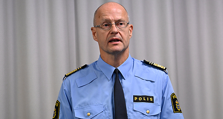 Han står och pratar med journalister. Han har poliskläder på sig.