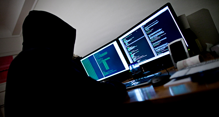 En person med luva på huvudet sitter framför flera datorskärmar.