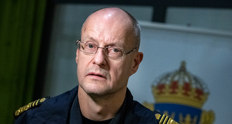 Han har glasögon och poliskläder på sig. Han tittar in i kameran.