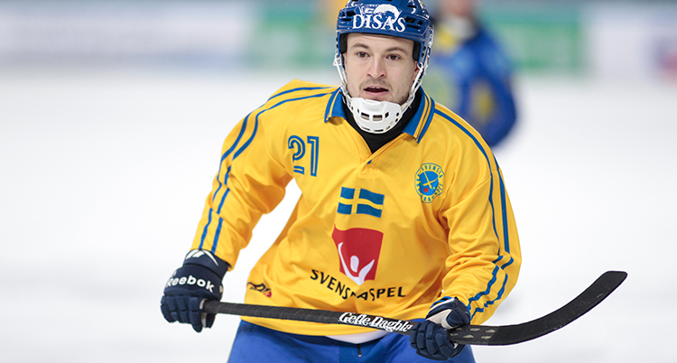 Närbild på en spelare under en match. Han har gul matchtröja och blå hjälm och håller i en bandyklubba.