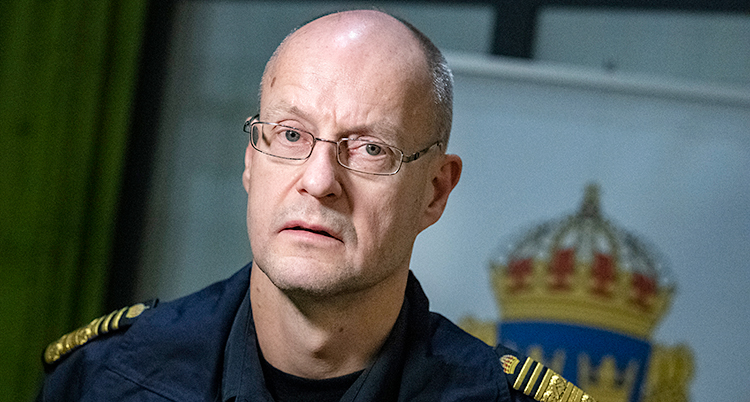 Han ser allvarlig ut. Han har tunt hår och glasögon och poliskläder.