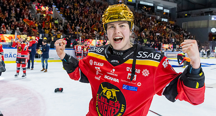 Hon är glad och knyter nävarna. Hon har på sig ishockey-kläder och står på isen.