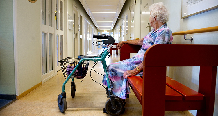 En äldre kvinna med rollator sitter på en bänk.