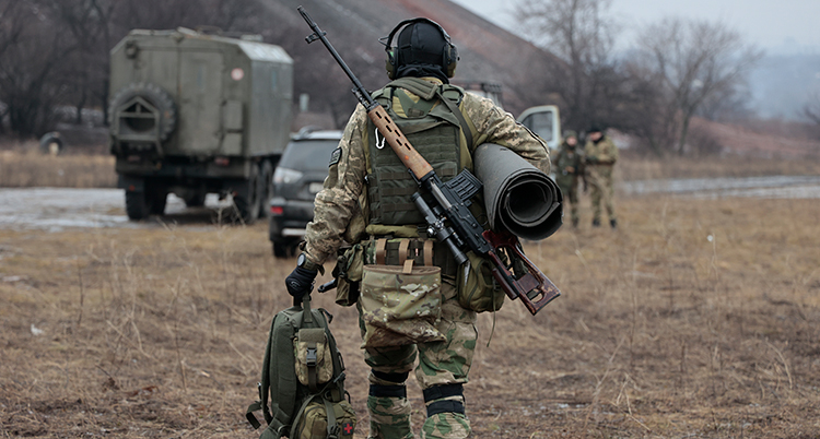 Ryggen på en soldat som går mot några bilar. Han har ett stort vapen på ryggen.