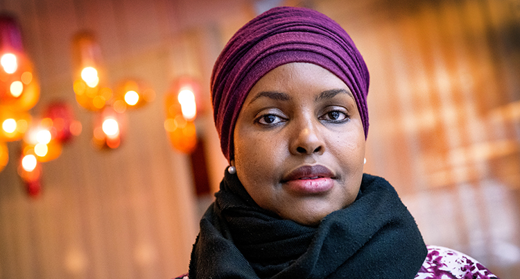 Det är ett nära porträtt av kvinnan. Hon har en lila sjal på huvudet, en svart sjal runt halsen. Hon tittar rakt in i kameran.