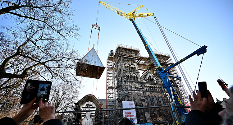 En stor kran lyfter ett torn upp mot kyrkan. Människor tar bilder med sina mobiler i förgrunden.