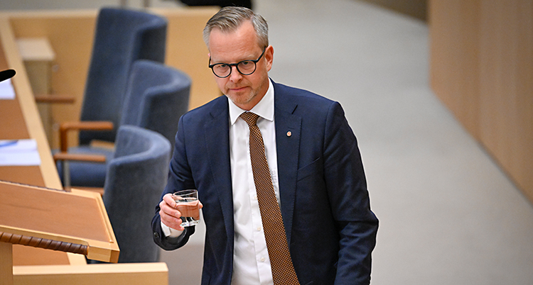 Han är i riksdagens stora sal. Han går med ett glas vatten i handen.