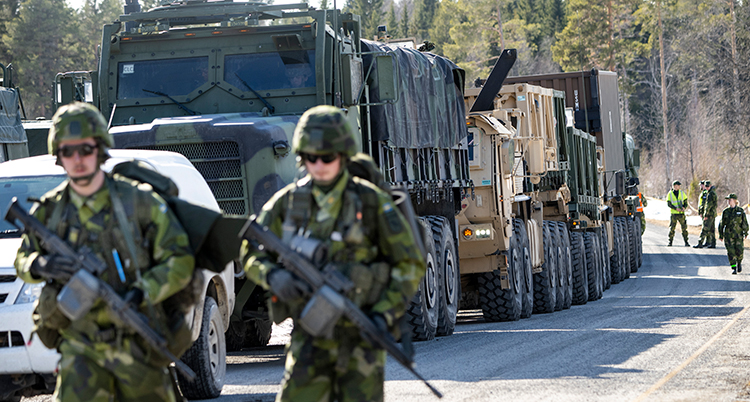Två militärer går längst fram. DE har vapen. Bakom syns många militärfordon på en väg. Flera militärer går på vägen.