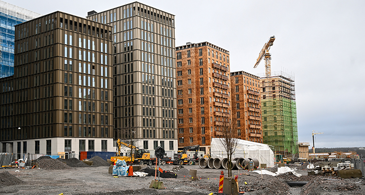 Flera höga hus syns. Framför är det grus på marken och byggmaterial. En kran syns till höger i bilden.