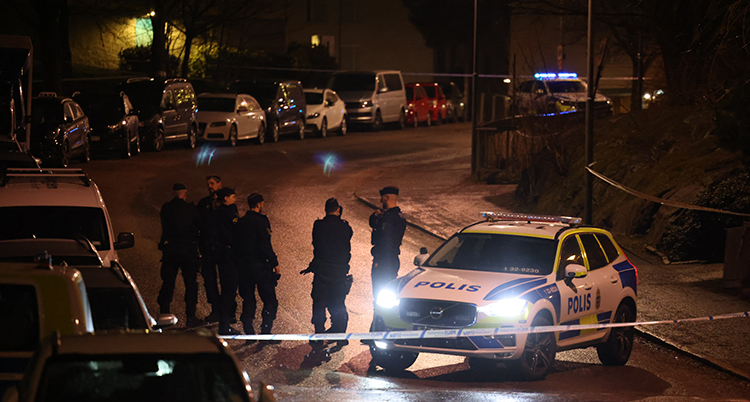 En polisbil står mitt i vägen. Poliser står runtomkring. Det är mörkt ute. Bilar står parkerade längs kanten av vägen. I bakgrunden syns hus.