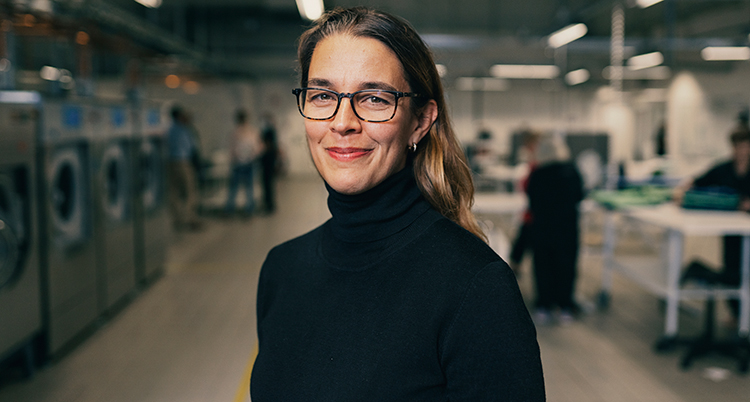 Hon tittar in i kameran. Hon har svart tröja och glasögon. Bakom syns tvättmaskiner och folk som jobbar vid bänkar.