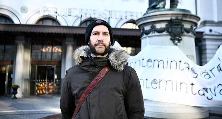 Han har jacka och mössa och skägg. Han står framför centralstationen.