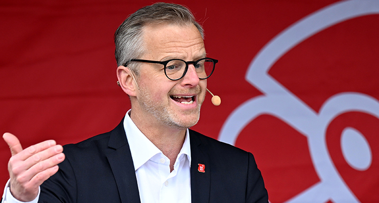 Han talar. Han har en mikrofon vid munnen. Bakgrunden är röd.