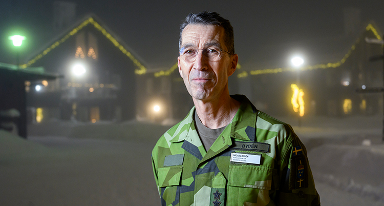 Han har gröna militärkläder. Han står utomhus. Han tittar in i kameran.