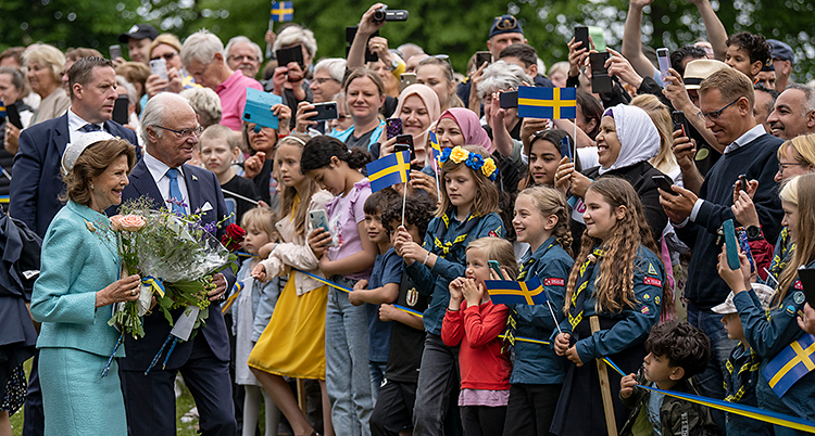 kungen och drottningen framför massor av människor med flaggor och blommor.
