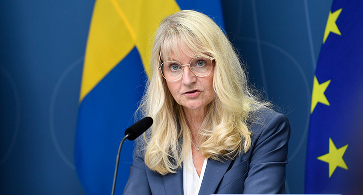 Hon har långt ljust hår och glasögon. Hon pratar i en mikrofon. Bakom syns flaggor.