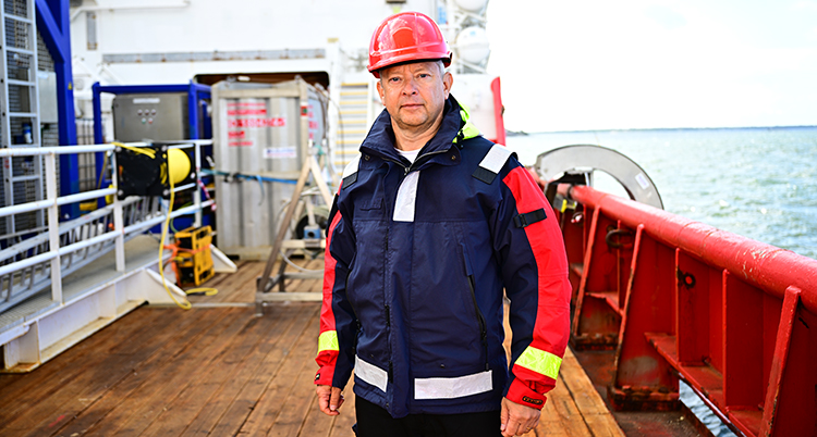 Han står på däck på en båt. Han har blå jacka och en röd hjälm. Havet syns bakom.