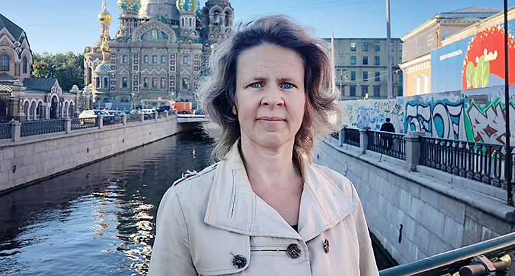 Hon står vid en flod i en stad. Hon tittar in i kameran.