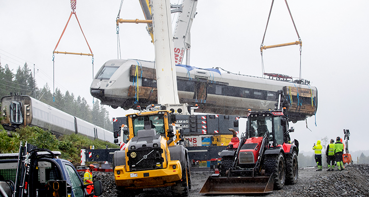 Traktorer står på vägen. Bakom lyfts den stora tågvagnen i luften med en stor kranarm.