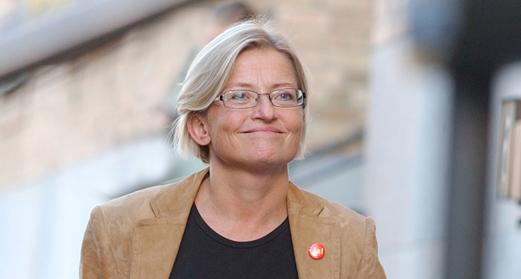 Ett porträtt fotograferat utomhus med ett hus i bakgrunden. Hon ler, har ljust hår och glasögon.