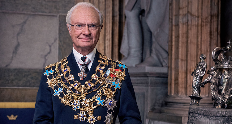 Han tittar in i kameran. Han bär många medaljer och halsband ovanpå kostymen.