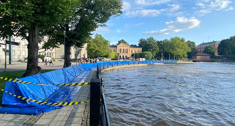 En blå mur av plast är byggd intill kanten av ån. Träd och hus finns vid kanten.