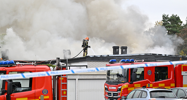 En brandman står på ett tak och sprutar vatten. Nedanför står flera brandbilar.