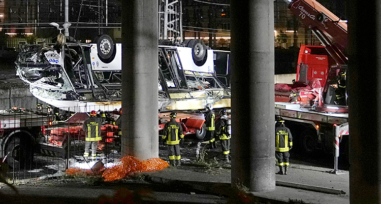 Flera brandmän står vid en vit buss. Bussen har ramlat ner från en bro och hamnat upp och ner. Den är mycket trasig.