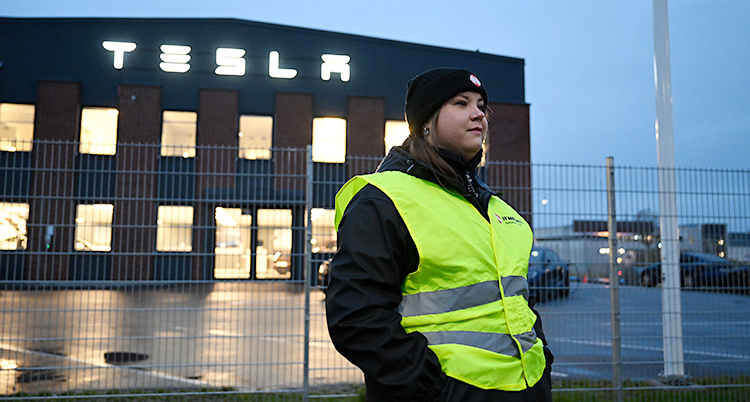 Hon står utanför Teslas verkstad. Hon har mössa och varselväst.
