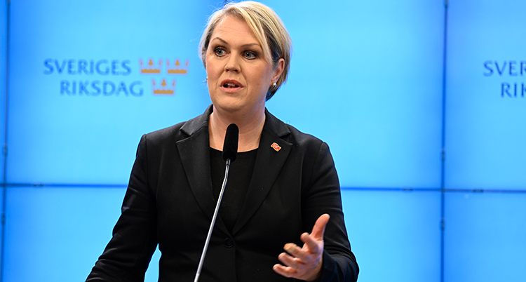 Hon pratar i en mikrofon. Hon har svarta kläde roch ljust hår. Bakgrunden är ljusblå och det står Sveriges Riksdag på väggen.