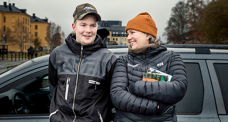 En ung kille och en kvinna står bredvid varandra framför en bil. De är glada.