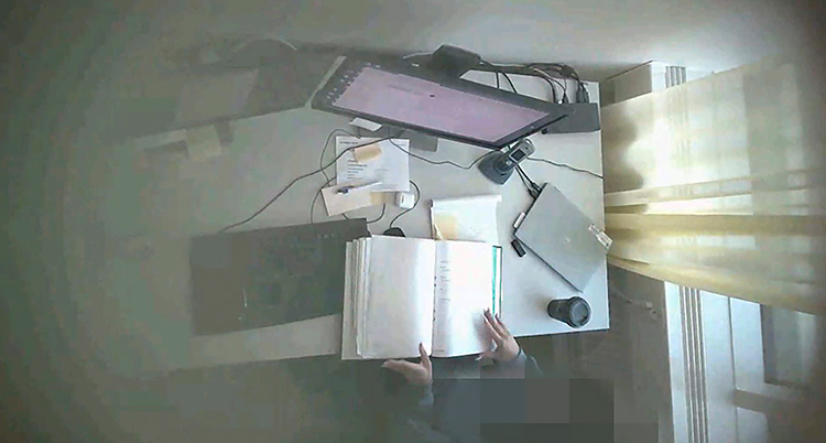 En suddig bild tagen ovanifrån på en person vid ett skrivbord. Vi ser bara händerna och allt på skrivbordet.