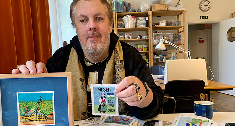 Han sitter vid ett bord. Han håller upp två broderade bilder.