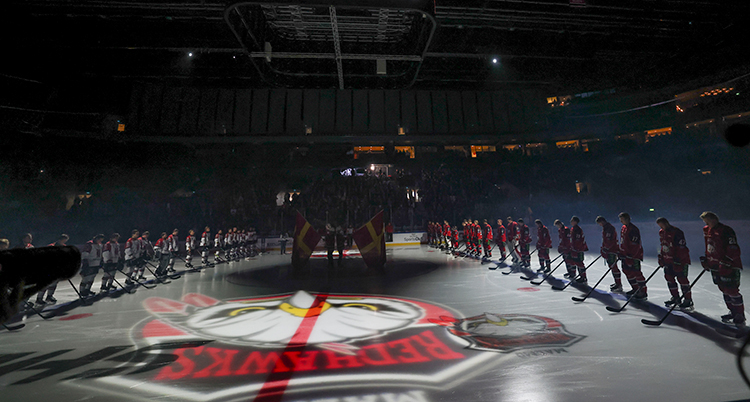 Två rader med spelare står stilla på isen i en nedsläckt arena.